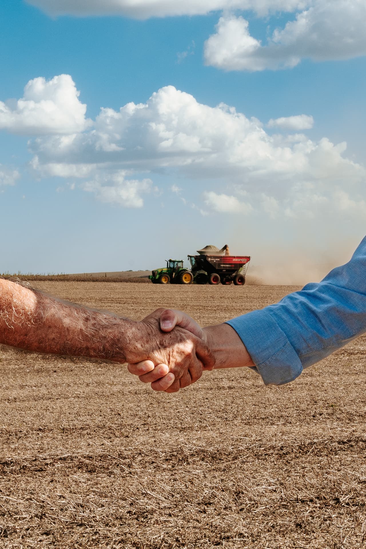 Aperto de mão entre produtor e consultor no campo, com operação agrícola ao fundo